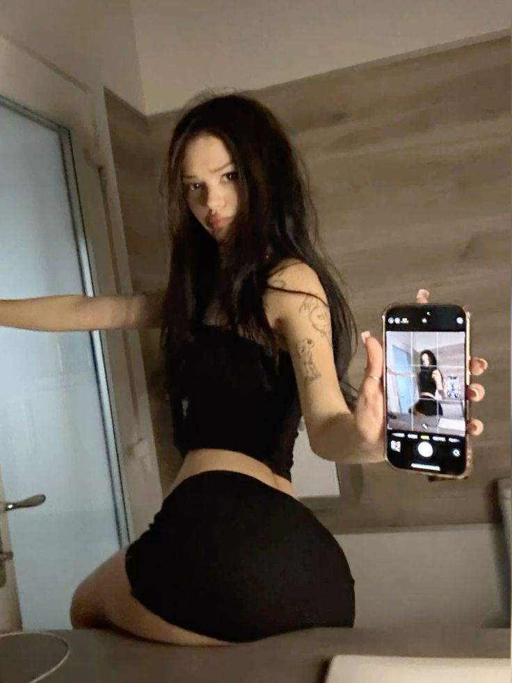 Woman in black top and skirt, holding phone showing her picture