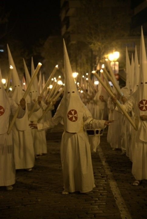 A group of people in white, pointed hoods and robes, holding candles, in a procession at night