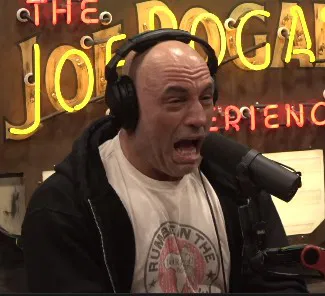 Joe Rogan wearing headphones and a t-shirt, with his mouth open in a surprised or yelling expression, in front of a neon sign