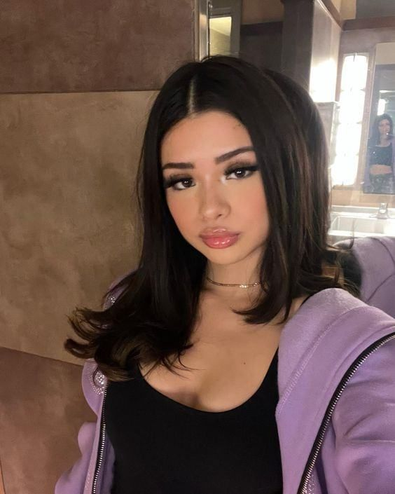A selfie of a young woman with long dark hair, wearing a black top and a light purple zip-up hoodie, looking at the camera