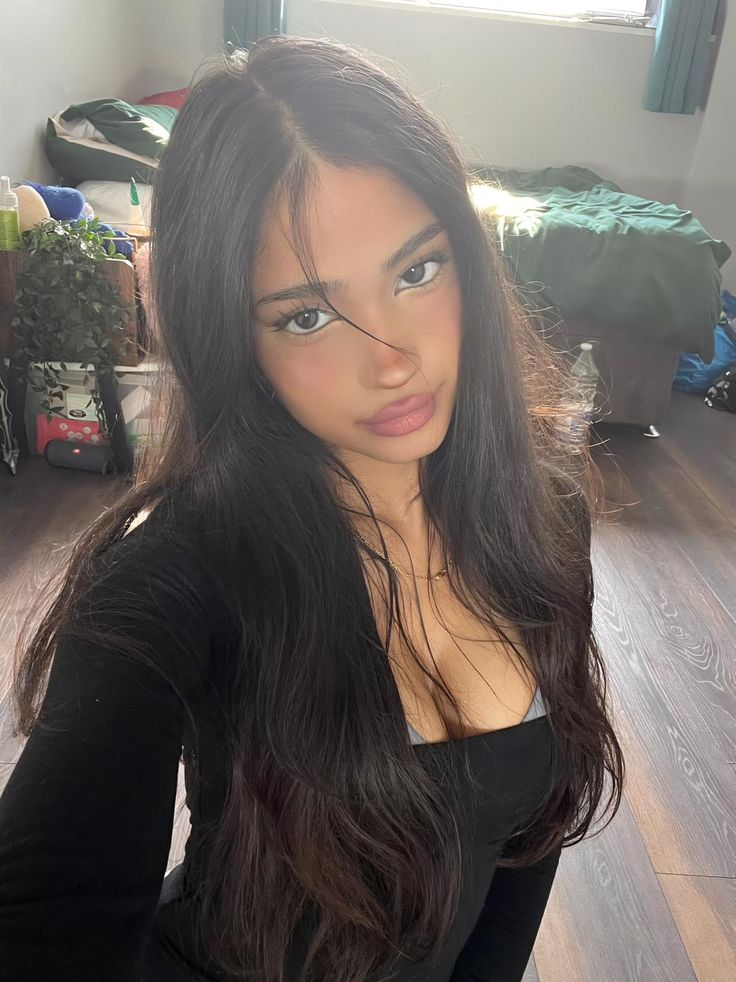 A selfie of a young woman with long dark hair, wearing a black long-sleeve top, standing in a room with wooden floors