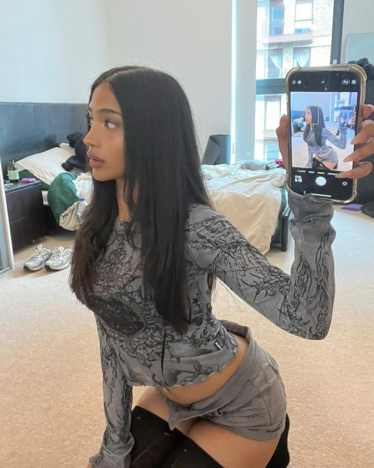 A young woman with long dark hair, wearing a grey patterned long-sleeve top and shorts, kneeling and holding up a phone to take a mirror selfie