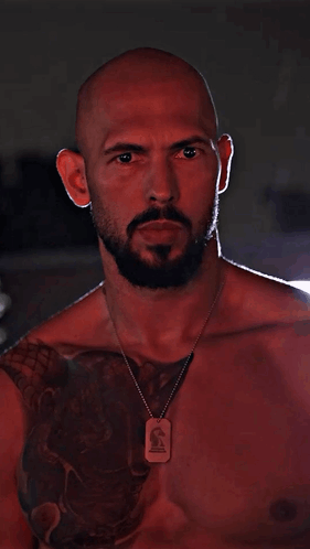 A bald man with a beard and a dog tag necklace, illuminated by red light, looking intently at the camera
