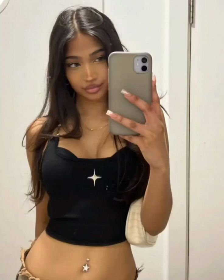 A selfie of a young woman with long dark hair, wearing a black tank top with a star design and a belly button piercing, holding a phone
