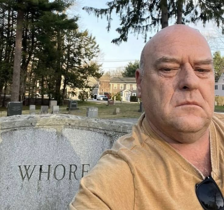 A bald man with a serious expression taking a selfie in a cemetery, with a tombstone in the foreground that has 'WHORE' carved into it