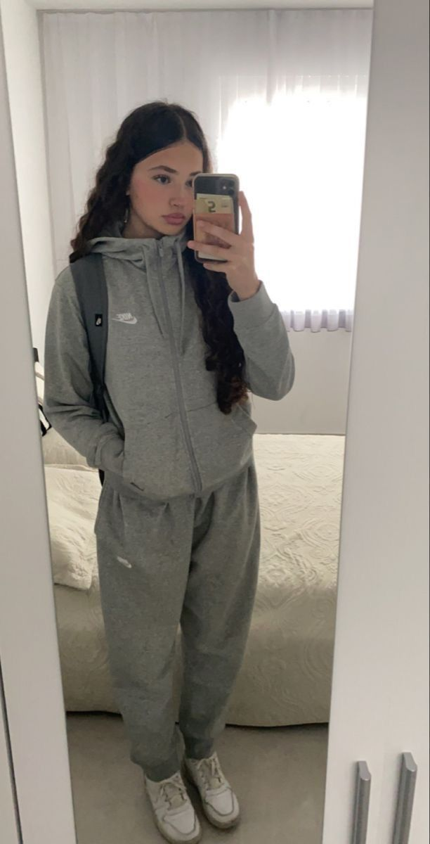 A young woman with long, curly dark hair, wearing a grey Nike tracksuit and white sneakers, taking a mirror selfie