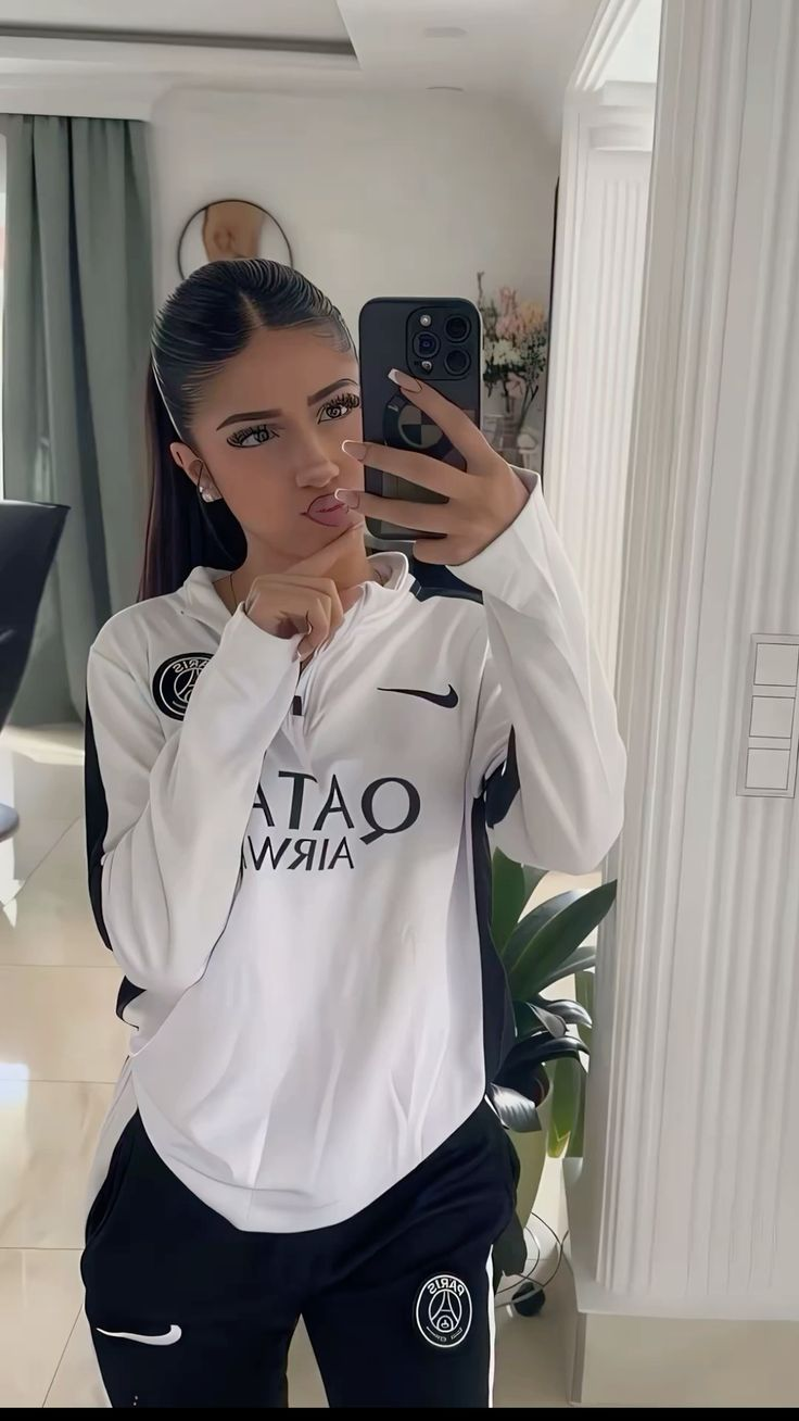 A young woman with dark hair tied in a high ponytail, wearing a white and black PSG (Paris Saint-Germain) half-zip top and black sweatpants, taking a mirror selfie
