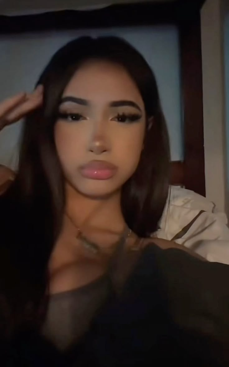 A close-up selfie of a young woman with long dark hair and full lips, wearing a light-colored top, looking at the camera