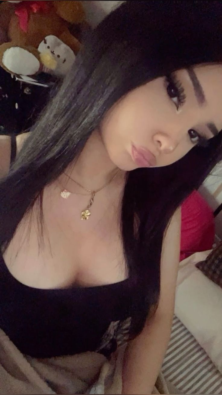 A selfie of a young woman with long dark hair, wearing a black tank top and gold necklaces, looking at the camera