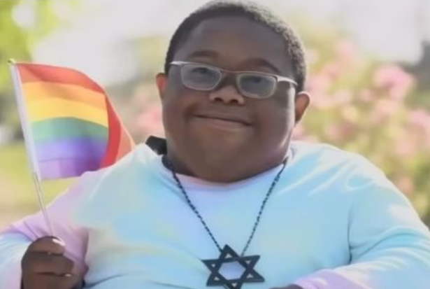 A young boy with glasses, wearing a light blue and pink tie-dye shirt and a Star of David necklace, holding a small rainbow flag