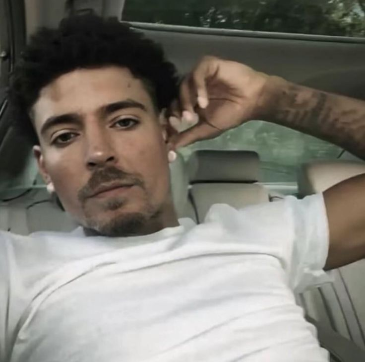 A man with curly dark hair and a beard, wearing a white t-shirt, sitting in a car and looking at the camera