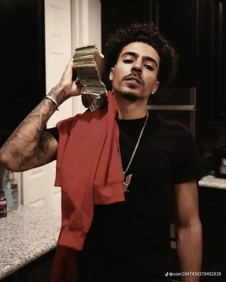 A man with curly dark hair and a beard, wearing a black t-shirt and a chain, holding a stack of money over his shoulder