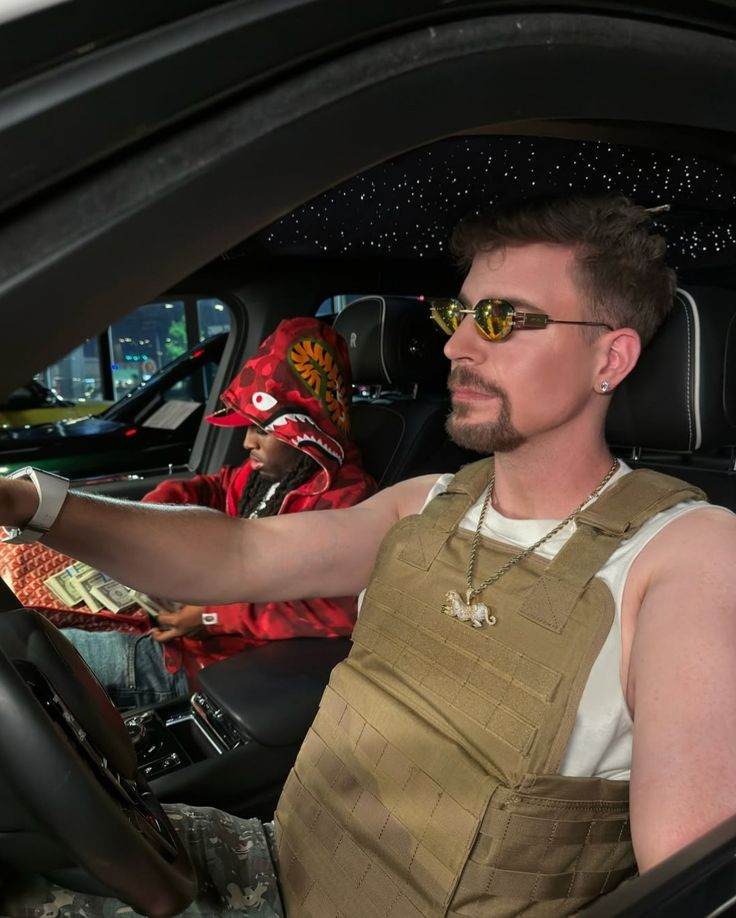 A man with a beard and sunglasses, wearing a white tank top and a tactical vest, driving a car with a starry ceiling, with another person in the passenger seat wearing a red hoodie