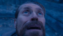 A close-up of Willem Dafoe looking upwards with a distressed expression, under blue lighting