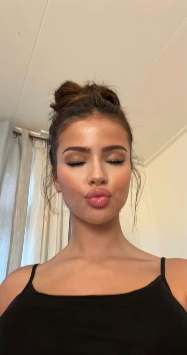 A close-up selfie of a young woman with her hair in a bun, eyes closed, making a kissy face, with prominent contouring