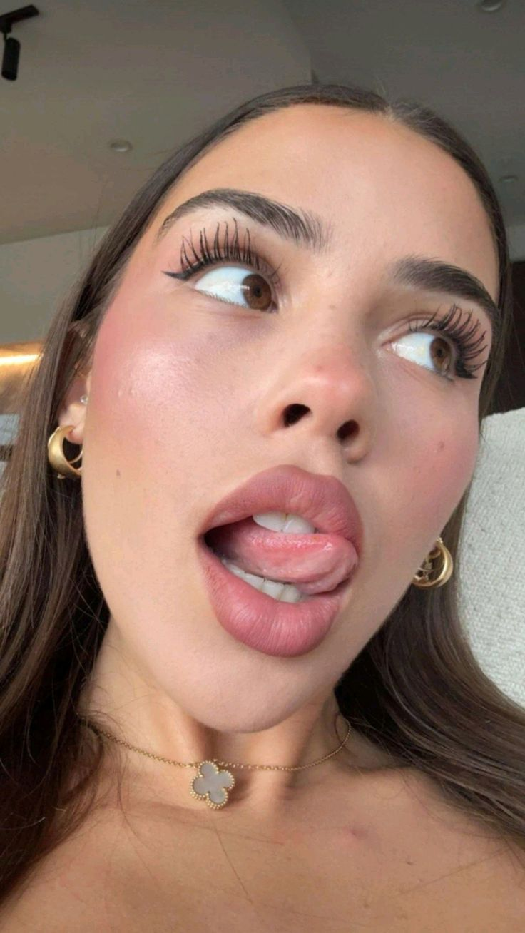 A close-up of a young woman's face, looking upwards and to the side, with her tongue slightly out, wearing gold hoop earrings and a clover necklace