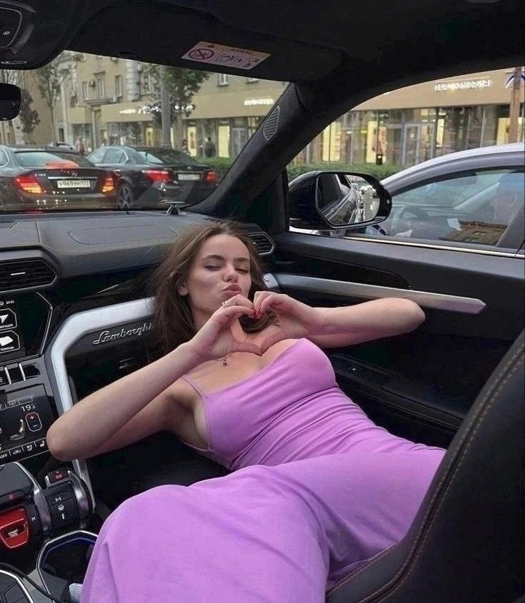 A young woman in a pink strapless dress sitting in a luxury car, making a heart shape with her hands